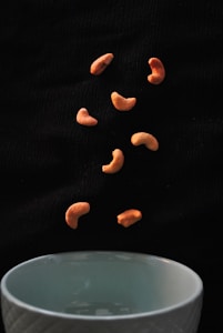 Several cashew nuts are captured in mid-air against a dark background, appearing to be falling towards a white bowl positioned at the bottom of the image.