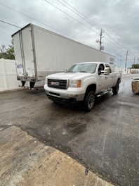 A white GMC pickup truck is parked next to a large white delivery trailer on a wet asphalt surface. The sky is overcast with gray clouds, and there are power lines visible overhead. A white fence runs along the side of the road and a small pile of cargo or debris can be seen to the right.