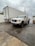 A white GMC pickup truck is parked next to a large white delivery trailer on a wet asphalt surface. The sky is overcast with gray clouds, and there are power lines visible overhead. A white fence runs along the side of the road and a small pile of cargo or debris can be seen to the right.