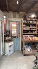 A rustic shop interior with wooden shelves filled with jars and packaged goods. A blue and white vintage door is adorned with a chalkboard sign advertising local free range eggs. The ceiling is wooden with an exposed bulb hanging. The floor is concrete, and one can see a refrigerator on the left and a decorated Christmas tree on the right.