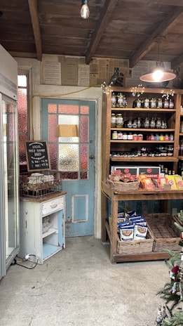 A rustic shop interior with wooden shelves filled with jars and packaged goods. A blue and white vintage door is adorned with a chalkboard sign advertising local free range eggs. The ceiling is wooden with an exposed bulb hanging. The floor is concrete, and one can see a refrigerator on the left and a decorated Christmas tree on the right.