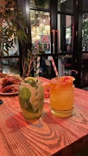 Two beverages sit on a wooden table in a cozy restaurant setting. One drink is clear with mint leaves and a striped straw, while the other is orange with a cherry garnish and a similar striped straw. In the background, there are plants, dim lighting, and diners at tables inside, adding a warm and inviting atmosphere.