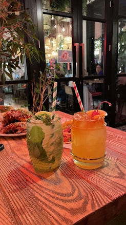 Two beverages sit on a wooden table in a cozy restaurant setting. One drink is clear with mint leaves and a striped straw, while the other is orange with a cherry garnish and a similar striped straw. In the background, there are plants, dim lighting, and diners at tables inside, adding a warm and inviting atmosphere.