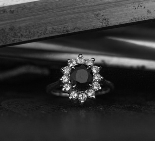 A close-up of an ornate ring featuring a central dark gemstone surrounded by smaller clear gemstones, set against a blurred metallic background.