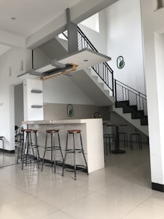 A modern, minimalistic interior space featuring a small bar with high stools and a sleek staircase with a metal railing. The color scheme is predominantly white and gray, with accents of light wood. The area is clean, well-lit with natural light, and has a contemporary design aesthetic.