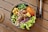 A bowl of mixed fresh ingredients including shredded carrots, edamame, diced mango, seaweed, cashews, green leafy vegetables, purple cabbage, and pieces of cooked fish. The bowl is placed on a wooden surface with a pair of silver utensils nearby.