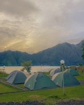 A campsite with several green tents is set on a grassy area beside a tranquil lake, surrounded by lush, green hills. The sky is filled with dramatic clouds, suggesting an early morning or late afternoon setting.