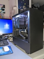 A desktop computer setup is visible with an open black computer case showing internal components such as a large cooling fan, cables, and a graphics card labeled 'GIGABYTE'. A monitor displaying the Windows login screen, a keyboard, and a mouse on a blue mouse pad are also present. On the wall, there are several posters featuring anime characters and a small lamp. The environment looks organized and clean.