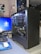 A desktop computer setup is visible with an open black computer case showing internal components such as a large cooling fan, cables, and a graphics card labeled 'GIGABYTE'. A monitor displaying the Windows login screen, a keyboard, and a mouse on a blue mouse pad are also present. On the wall, there are several posters featuring anime characters and a small lamp. The environment looks organized and clean.