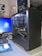 A desktop computer setup is visible with an open black computer case showing internal components such as a large cooling fan, cables, and a graphics card labeled 'GIGABYTE'. A monitor displaying the Windows login screen, a keyboard, and a mouse on a blue mouse pad are also present. On the wall, there are several posters featuring anime characters and a small lamp. The environment looks organized and clean.
