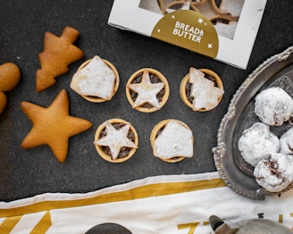 An assortment of baked goods, including star-shaped gingerbread cookies, pastry tarts with star-patterned toppings, and snow-dusted chocolate treats on a vintage silver tray. A partially visible box labeled 'Bread & Butter' is present, along with a decorative cloth.