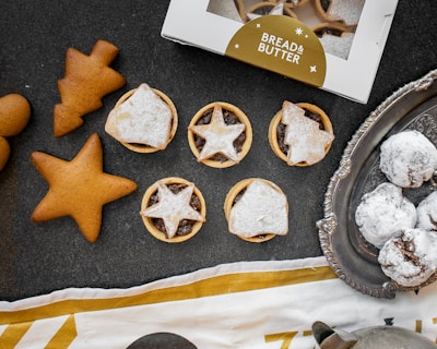 An assortment of baked goods, including star-shaped gingerbread cookies, pastry tarts with star-patterned toppings, and snow-dusted chocolate treats on a vintage silver tray. A partially visible box labeled 'Bread & Butter' is present, along with a decorative cloth.
