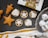 An assortment of baked goods, including star-shaped gingerbread cookies, pastry tarts with star-patterned toppings, and snow-dusted chocolate treats on a vintage silver tray. A partially visible box labeled 'Bread & Butter' is present, along with a decorative cloth.