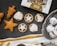 An assortment of baked goods, including star-shaped gingerbread cookies, pastry tarts with star-patterned toppings, and snow-dusted chocolate treats on a vintage silver tray. A partially visible box labeled 'Bread & Butter' is present, along with a decorative cloth.
