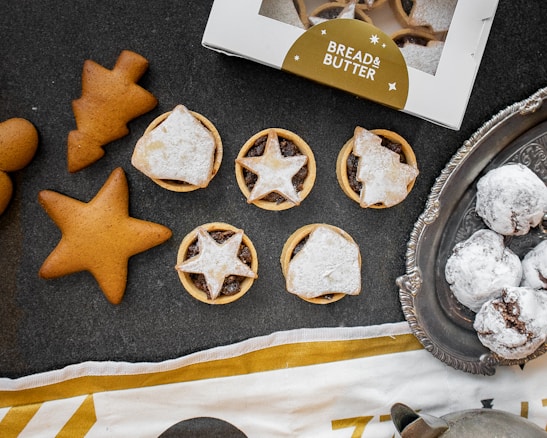 An assortment of baked goods, including star-shaped gingerbread cookies, pastry tarts with star-patterned toppings, and snow-dusted chocolate treats on a vintage silver tray. A partially visible box labeled 'Bread & Butter' is present, along with a decorative cloth.