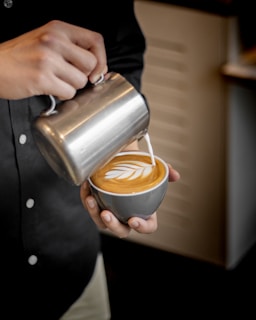 A person is skillfully pouring steamed milk from a metal jug into a coffee cup to create latte art, illustrating a delicate leaf pattern on the coffee's surface. The person is wearing a dark shirt with visible buttons.