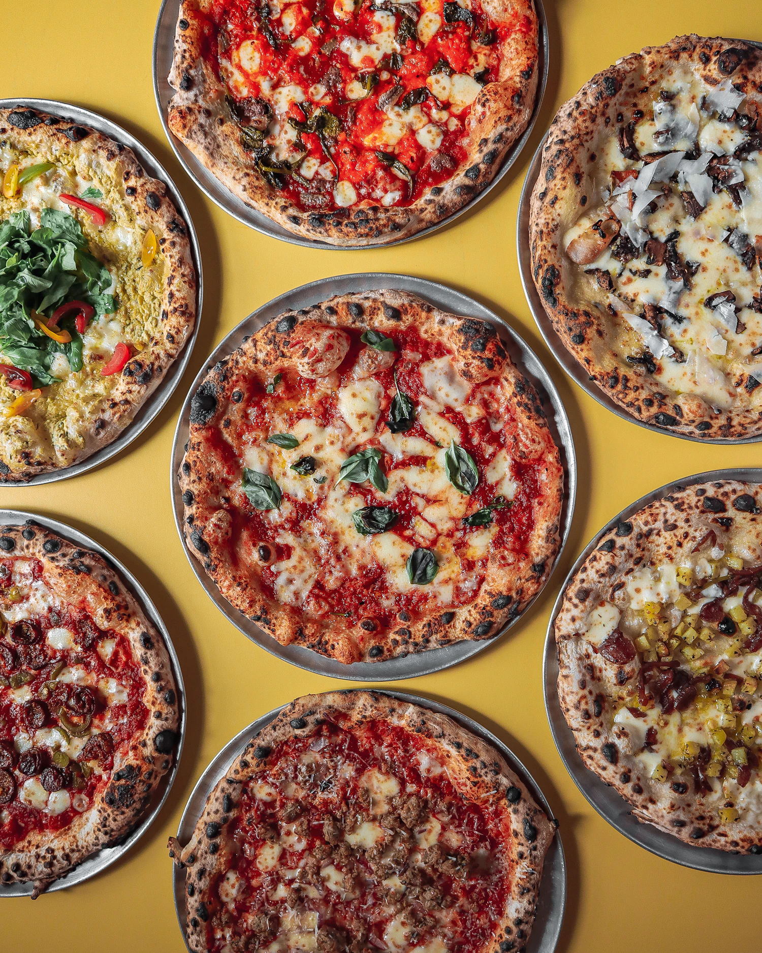 Six whole pizzas laid out side by side on a yellow surface