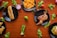 A variety of foods arranged on a wooden table, including a hot dog with mustard, French fries, a roasted chicken, samosas, a burger, and scattered lettuce leaves. Each dish is placed on a black plate, surrounded by assorted vegetables like cucumbers, tomatoes, and lime.