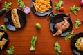 A variety of foods arranged on a wooden table, including a hot dog with mustard, French fries, a roasted chicken, samosas, a burger, and scattered lettuce leaves. Each dish is placed on a black plate, surrounded by assorted vegetables like cucumbers, tomatoes, and lime.