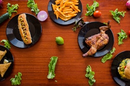 A variety of foods arranged on a wooden table, including a hot dog with mustard, French fries, a roasted chicken, samosas, a burger, and scattered lettuce leaves. Each dish is placed on a black plate, surrounded by assorted vegetables like cucumbers, tomatoes, and lime.