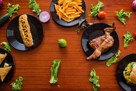 A variety of foods arranged on a wooden table, including a hot dog with mustard, French fries, a roasted chicken, samosas, a burger, and scattered lettuce leaves. Each dish is placed on a black plate, surrounded by assorted vegetables like cucumbers, tomatoes, and lime.