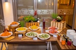 A well-organized buffet table displaying a variety of salads and condiments. There are bowls filled with different types of salads on wooden risers, decorated with fresh greens and flowers. Alongside the salads, there are jars of colorful sauces neatly arranged near a stack of white plates.
