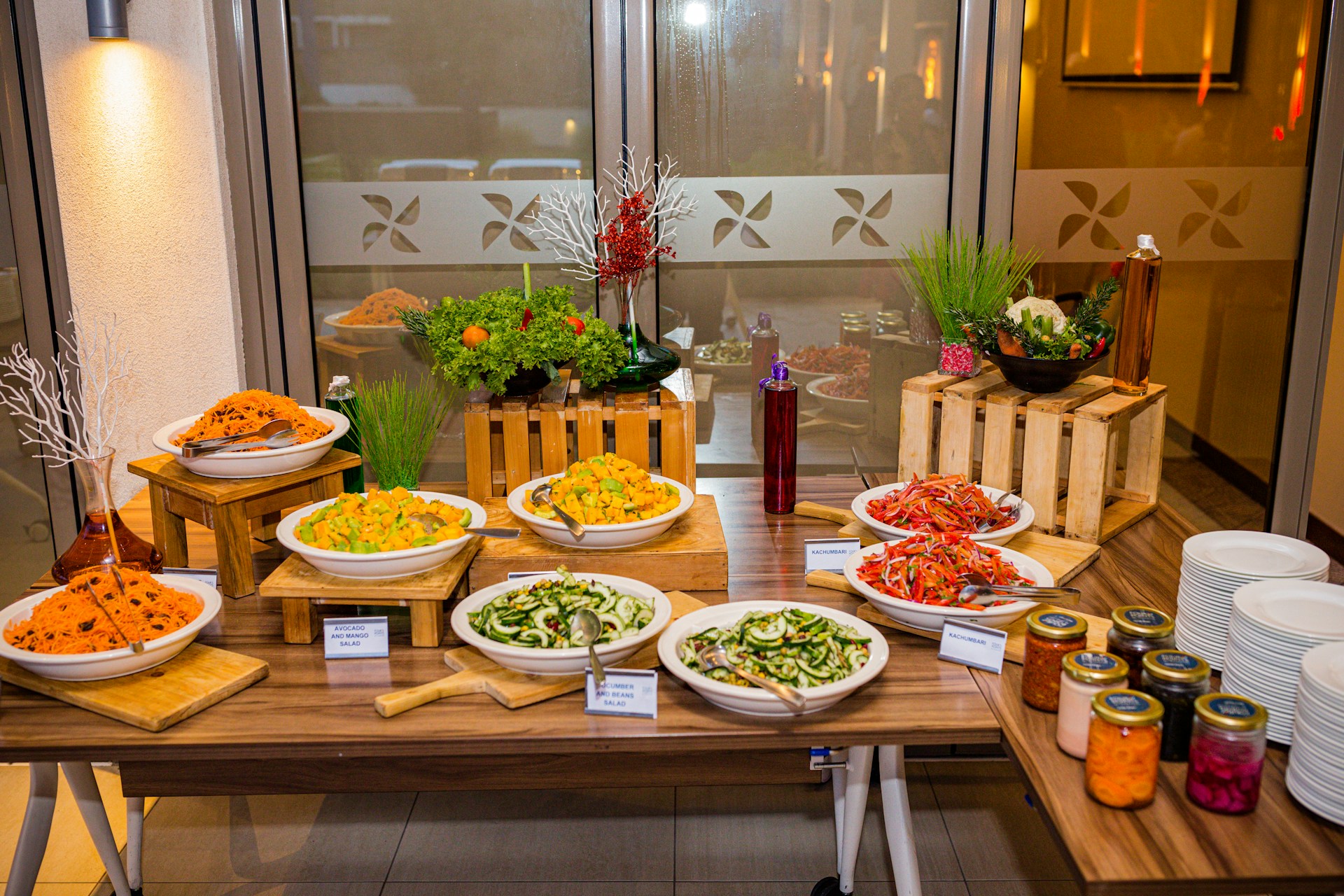 A well-organized buffet table displaying a variety of salads and condiments. There are bowls filled with different types of salads on wooden risers, decorated with fresh greens and flowers. Alongside the salads, there are jars of colorful sauces neatly arranged near a stack of white plates.