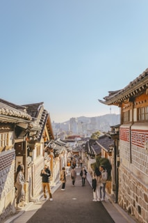 A traditional street with historic Korean architecture, featuring tiled roofs and brick walls. People walk along the street, some are taking photographs. In the background, a modern cityscape with tall buildings is visible under a clear blue sky.