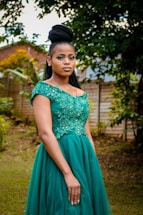 A woman in an elegant green dress stands outdoors. The dress features intricate floral embellishments on the bodice. Her hair is styled in a high braided updo. She is positioned in a garden-like setting with green foliage and a wooden fence in the background.
