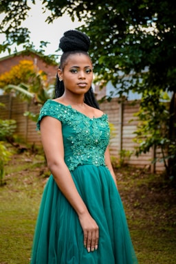 A woman in an elegant green dress stands outdoors. The dress features intricate floral embellishments on the bodice. Her hair is styled in a high braided updo. She is positioned in a garden-like setting with green foliage and a wooden fence in the background.