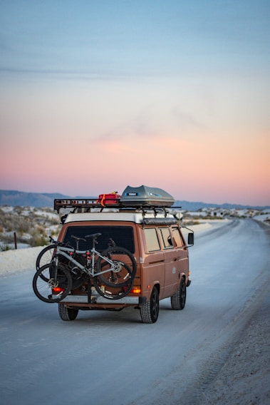 A vintage brown van travels down an empty, scenic road at dusk. Two bicycles are mounted on the back, and gear is stored on the roof rack.