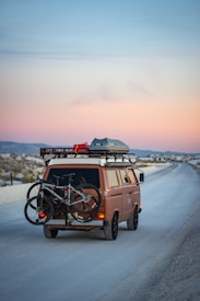 A vintage brown van travels down an empty, scenic road at dusk. Two bicycles are mounted on the back, and gear is stored on the roof rack.