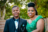 A young man and woman are dressed in formal attire outdoors. The man is wearing a black tuxedo with a bow tie, while the woman is in a vibrant green dress adorned with floral details. The background features greenery and an out-of-focus natural setting, indicating an outdoor event or celebration.