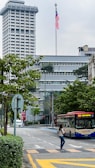 A city street scene featuring a tall building with a large flagpole displaying the Malaysian flag. In the foreground, a public bus labeled 'rapidKL' is on the road, with a woman crossing the street while looking at her phone. Lush green trees line the sidewalk, and several modern structures are visible in the background.