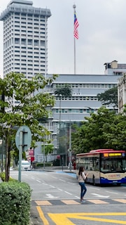 A city street scene featuring a tall building with a large flagpole displaying the Malaysian flag. In the foreground, a public bus labeled 'rapidKL' is on the road, with a woman crossing the street while looking at her phone. Lush green trees line the sidewalk, and several modern structures are visible in the background.