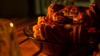 A glass dish contains several cooked crabs, their shells a rich reddish-orange, sitting on a wooden table. The lighting is warm and creates an intimate, cozy atmosphere, highlighting the textures and details of the crab shells.