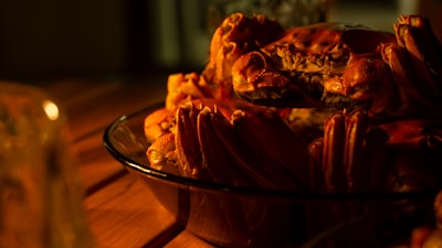 A glass dish contains several cooked crabs, their shells a rich reddish-orange, sitting on a wooden table. The lighting is warm and creates an intimate, cozy atmosphere, highlighting the textures and details of the crab shells.