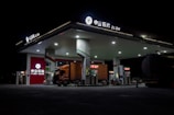 A gas station brightly lit at night with a large truck parked at the fuel pumps. The architecture is modern with a flat roof and multiple fuel dispensers. The signage at the top and on the building includes both Chinese characters and English letters. The station is relatively quiet and appears to be well-maintained, with the focus on the truck and the illuminated stations.