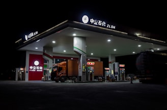 A gas station brightly lit at night with a large truck parked at the fuel pumps. The architecture is modern with a flat roof and multiple fuel dispensers. The signage at the top and on the building includes both Chinese characters and English letters. The station is relatively quiet and appears to be well-maintained, with the focus on the truck and the illuminated stations.