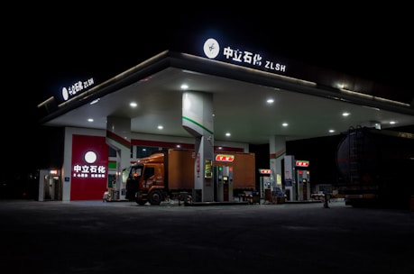 A gas station brightly lit at night with a large truck parked at the fuel pumps. The architecture is modern with a flat roof and multiple fuel dispensers. The signage at the top and on the building includes both Chinese characters and English letters. The station is relatively quiet and appears to be well-maintained, with the focus on the truck and the illuminated stations.