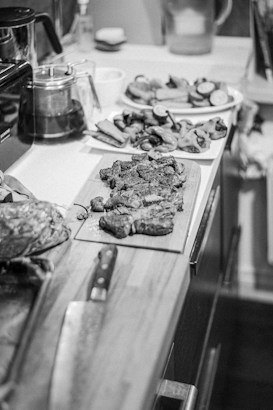 A black and white image of a kitchen counter featuring a cutting board with cooked meat, a large knife, and several plates of sliced vegetables. The countertop also has a coffee maker, a jar, and other small containers in the background.
