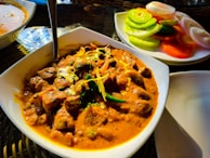 A dish of rich, creamy curry with chunks of meat, garnished with herbs and spices. Accompanying the curry is a side plate of sliced cucumbers, tomatoes, and onions, adding a fresh contrast. The setting suggests an appetizing dining experience.