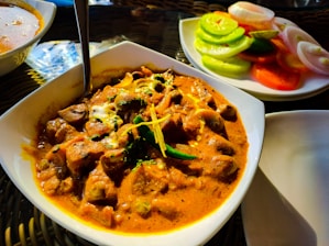 A dish of rich, creamy curry with chunks of meat, garnished with herbs and spices. Accompanying the curry is a side plate of sliced cucumbers, tomatoes, and onions, adding a fresh contrast. The setting suggests an appetizing dining experience.