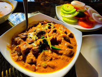 A dish of rich, creamy curry with chunks of meat, garnished with herbs and spices. Accompanying the curry is a side plate of sliced cucumbers, tomatoes, and onions, adding a fresh contrast. The setting suggests an appetizing dining experience.