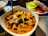 A dish of rich, creamy curry with chunks of meat, garnished with herbs and spices. Accompanying the curry is a side plate of sliced cucumbers, tomatoes, and onions, adding a fresh contrast. The setting suggests an appetizing dining experience.
