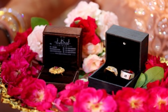 Two jewelry boxes are surrounded by vibrant red and pink flowers. The left box contains a gold ring with a red gemstone, while the right box holds two rings, one with a prominent gold band and the other with silver and red accents. The scene is decorated with soft petals, creating an elegant and luxurious setting.