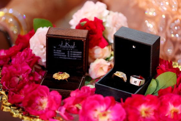 Two jewelry boxes are surrounded by vibrant red and pink flowers. The left box contains a gold ring with a red gemstone, while the right box holds two rings, one with a prominent gold band and the other with silver and red accents. The scene is decorated with soft petals, creating an elegant and luxurious setting.