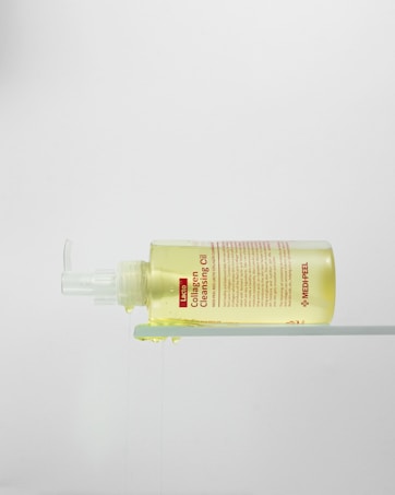 A bottle of Collagen Cleansing Oil with a pump dispenser is placed horizontally on a transparent surface, with the liquid slightly oozing out. The bottle is clear, revealing the yellowish oil inside. The label features product information in red and black text.