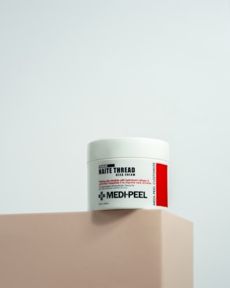 A jar of Naite Thread Neck Cream by MEDI-PEEL rests on a beige platform. The label displays information about the product, which includes enhancing skin elasticity with collagen and peptides. The background is a light, neutral shade which gives a minimalist and clean appearance.