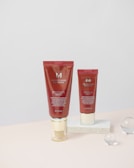 Two red cosmetic tubes labeled as BB cream are positioned vertically on a light-colored surface. Both tubes are marked with the brand name 'MISSHA' and they contain detailed product information. One tube is directly on the surface, while the other is placed on a white rectangular block. Clear glass spheres are scattered in the background, adding an aesthetic touch.